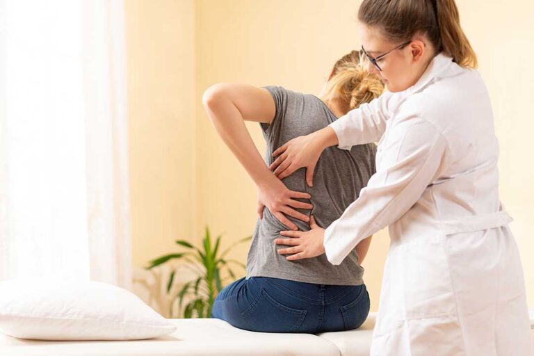 A patient discusses back pain with a clinician beside an examination table and anatomy chart.